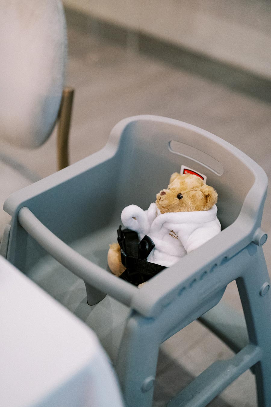 A plush teddy bear sits in a gray high chair, wearing a white robe with black straps, inside a dining area.