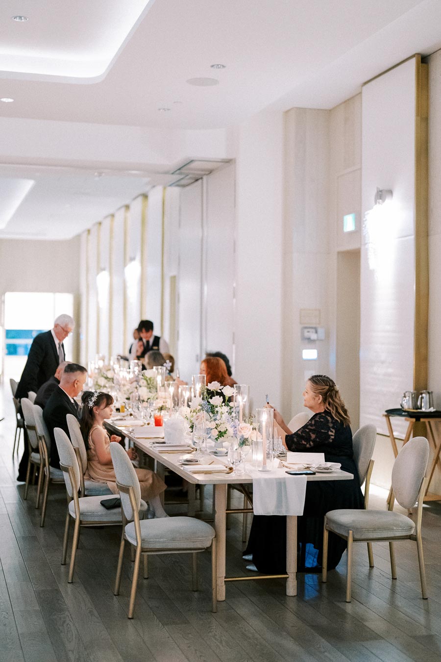Elegant wedding reception in a modern venue with guests seated at a long, beautifully decorated dining table adorned with floral arrangements and candles.