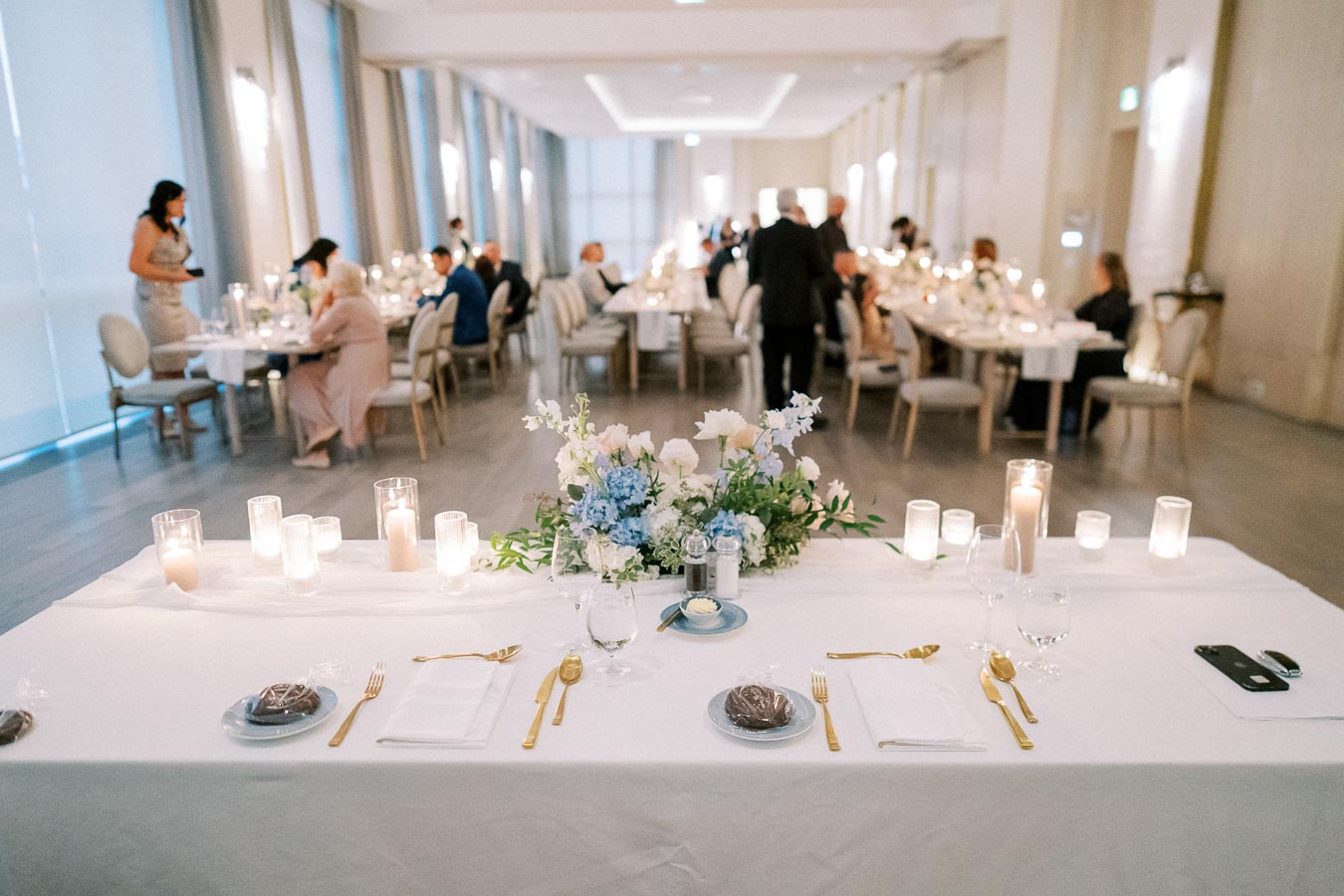 Elegant wedding reception hall with beautifully decorated tables featuring white linens, gold cutlery, floral centerpieces, and glowing candles, creating an inviting and romantic atmosphere.