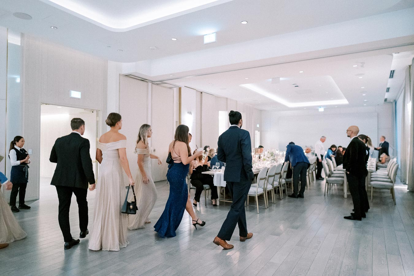 Elegant event attendees in formal attire walk towards a dining area in a sophisticated, well-lit banquet hall with neatly arranged tables and chairs.