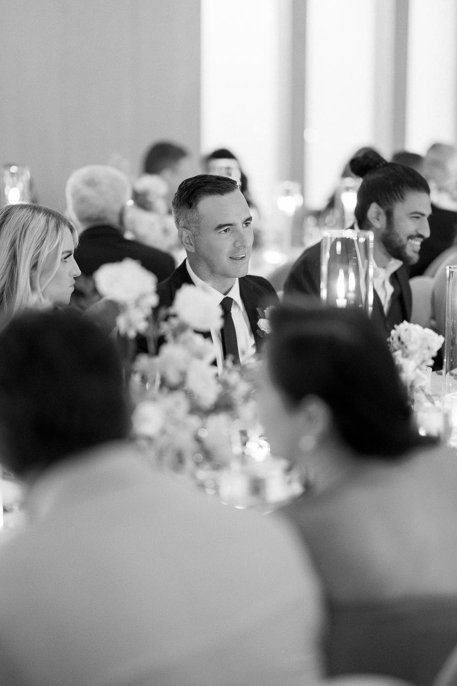 Black and white photo of a formal event with guests seated at a decorated table, engaged in conversation, with floral centerpieces and candlelit ambiance.