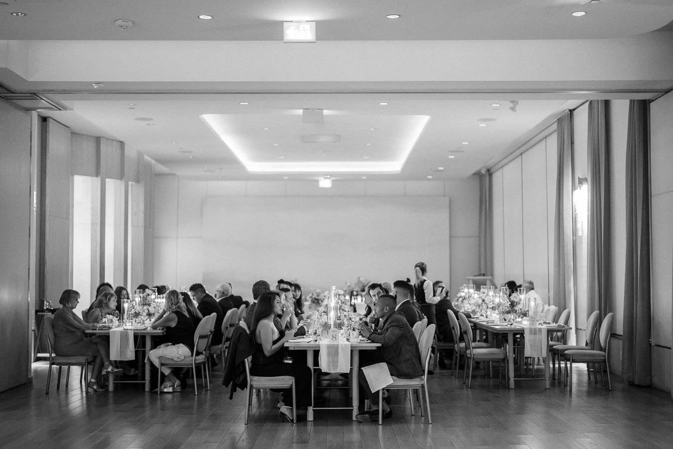 Elegant black and white photograph of a formal dining event, featuring guests seated at tables in a modern, well-lit banquet hall, with candlelit centerpieces and floor-to-ceiling curtains.