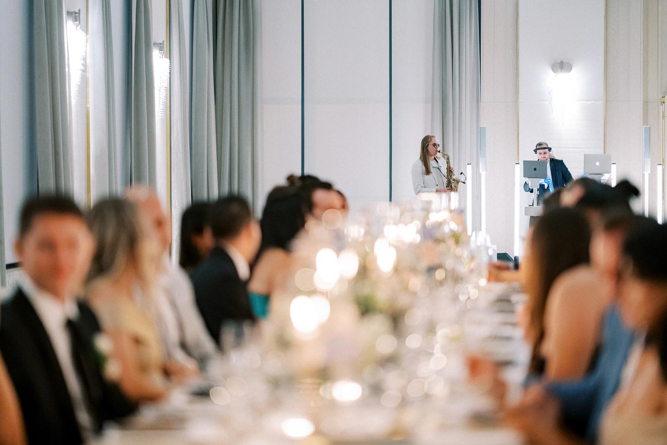 Guests seated at a formal dinner table with elegant place settings, while a speaker in the background performs with a saxophone, accompanied by a DJ at a modern indoor event.