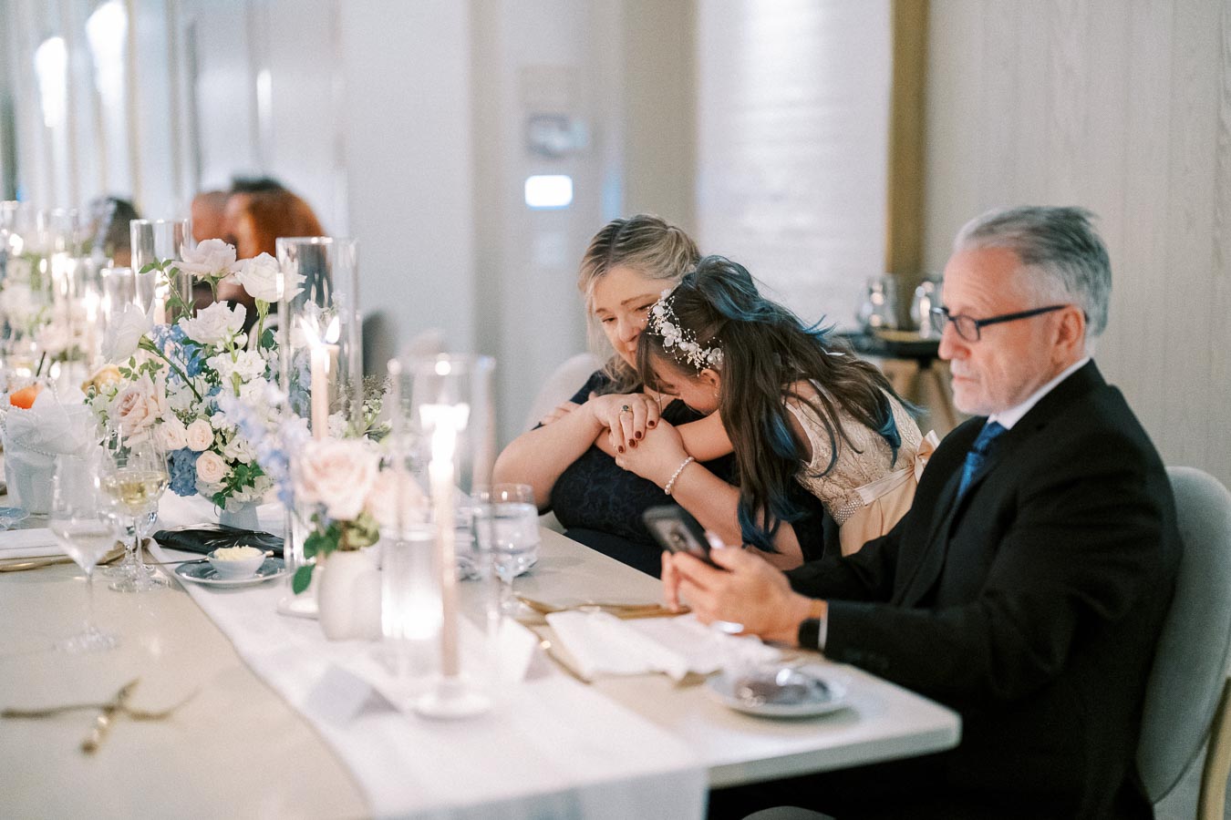 A beautifully decorated dinner table with elegant floral arrangements and glowing candles, where a man in a suit is seated next to two women sharing a heartfelt moment, creating a warm and intimate atmosphere at a formal event.