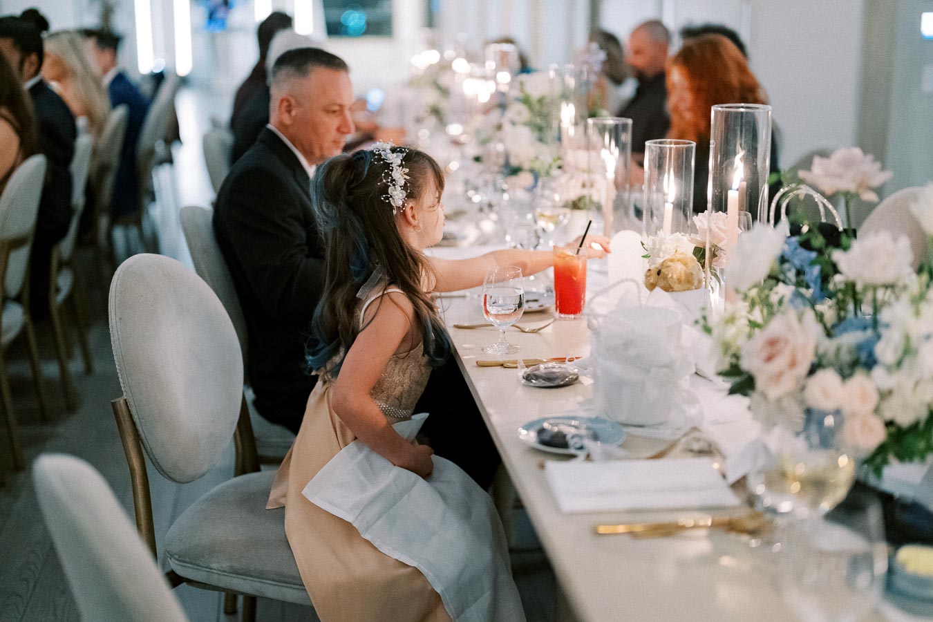 Elegant wedding reception table setting with floral centerpieces and candlelight, featuring guests enjoying a celebratory meal. A young girl in a fancy dress is seated beside an adult, creating a warm and festive atmosphere.
