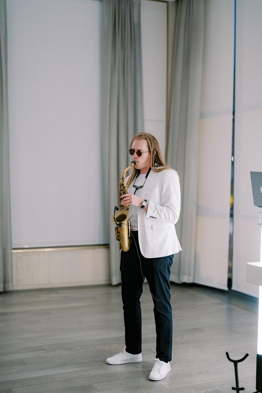 A musician with long hair plays a saxophone while wearing sunglasses and a white blazer. He stands on a light-colored wooden floor in a bright room with closed curtains.