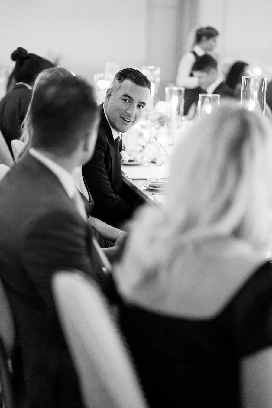 Black and white photo of a group of people dressed in formal attire, seated at a dinner table during an elegant event. A man in a suit looks attentively towards a person across the table. The atmosphere appears sophisticated and intimate, with soft lighting and decorative table settings.