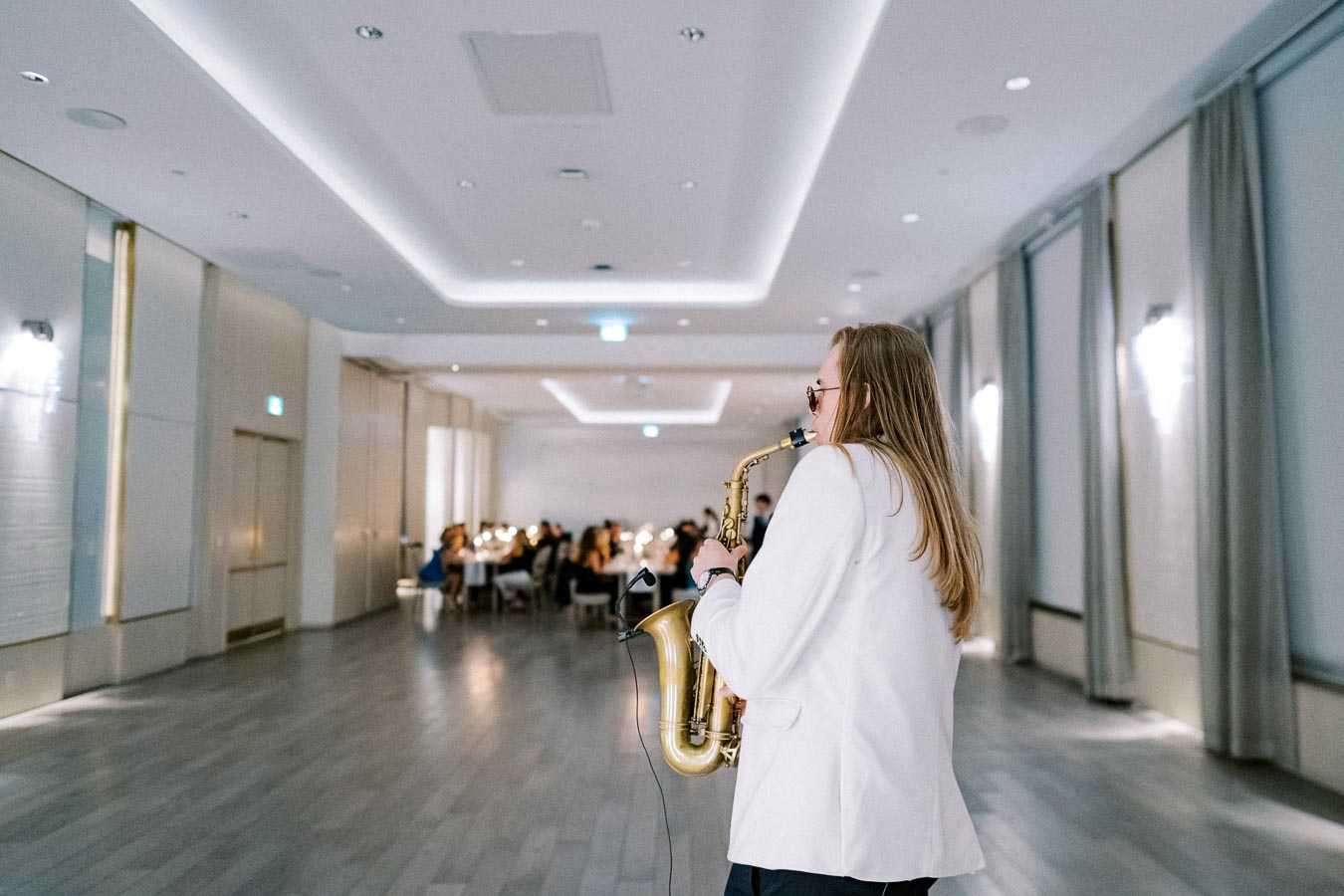 Saxophonist in a white suit playing at a formal event in a spacious, elegantly decorated room.