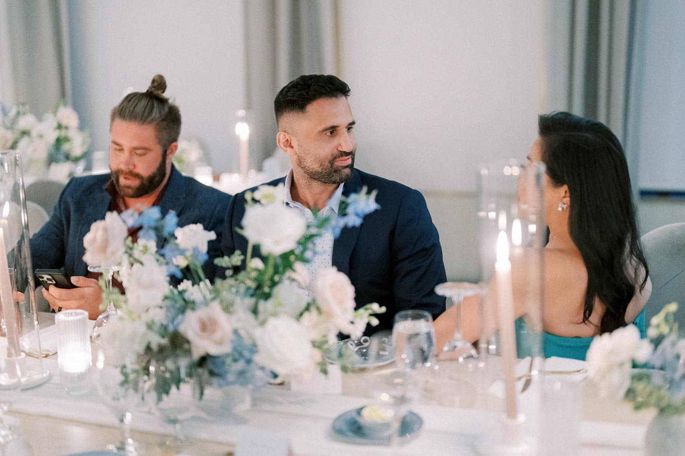 Three elegantly dressed people seated at a beautifully decorated dinner table adorned with white and blue flowers and lit candles, creating a sophisticated and intimate atmosphere.