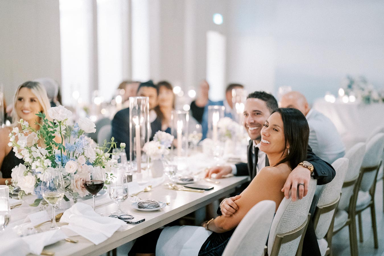 A joyful wedding reception scene with elegantly dressed guests seated at a long table adorned with beautiful white and blue floral arrangements and candlelight, creating a warm and festive atmosphere.