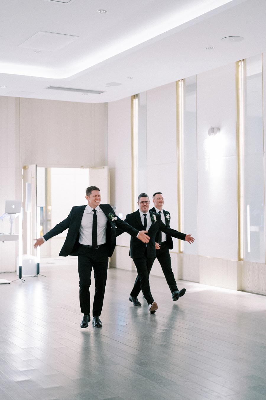 Three groomsmen in black suits and ties energetically enter a bright, modern wedding venue with smiles and open arms.