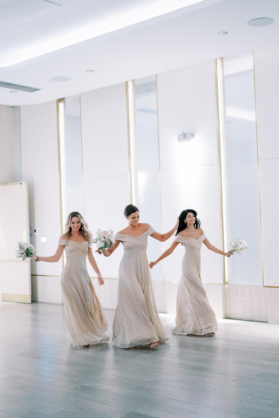 Three bridesmaids in elegant, off-shoulder champagne dresses joyfully walking and holding bouquets indoors, with a modern, minimalist background.