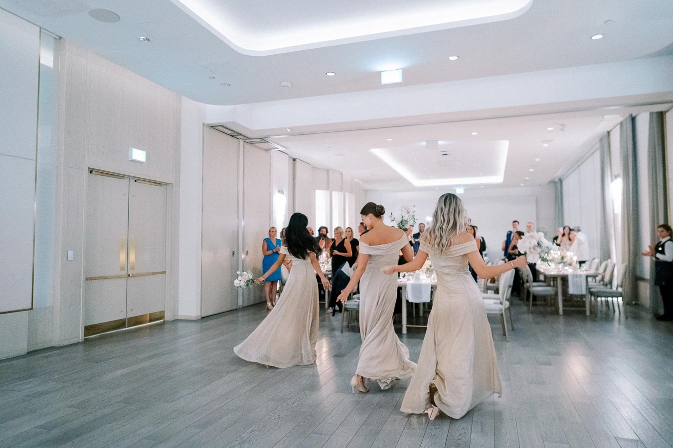 Bridesmaids in elegant beige dresses dance in a modern, well-lit reception hall with guests watching attentively.