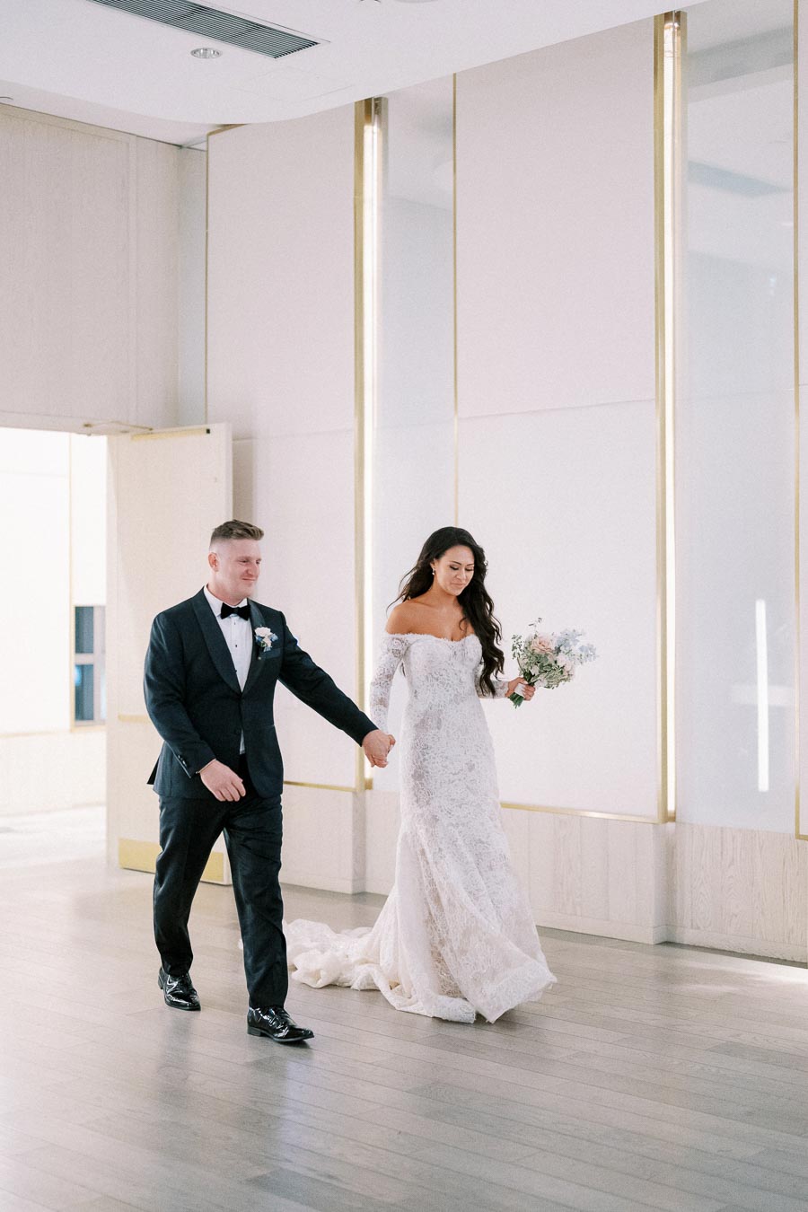 Newlywed couple walking hand in hand in elegant wedding attire, with bride in lace gown holding a bouquet, in a modern venue with light wood flooring.