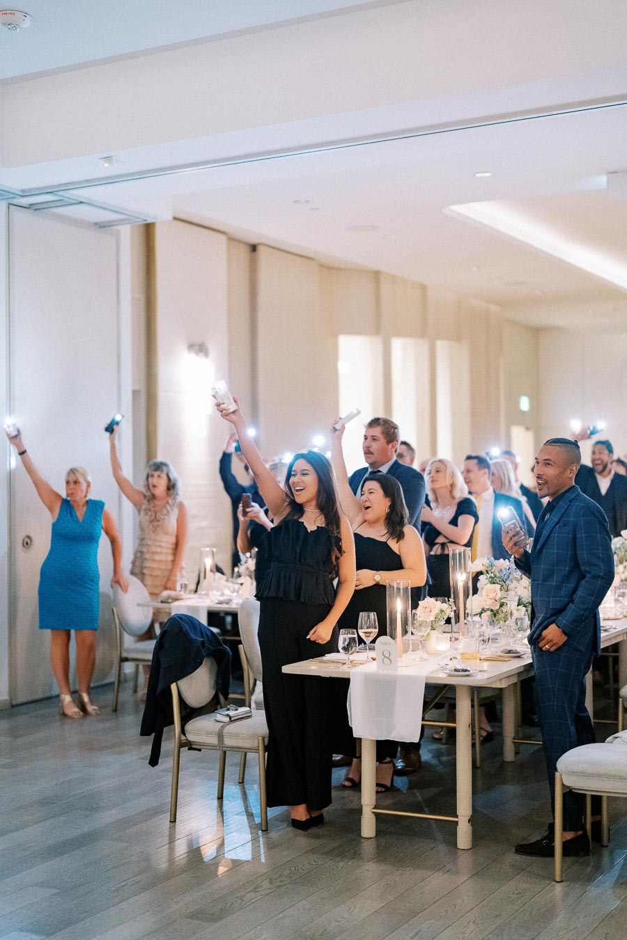 Guests joyfully raising their glasses and phone flashlights in celebration during a wedding reception, beautifully decorated tables in the foreground.
