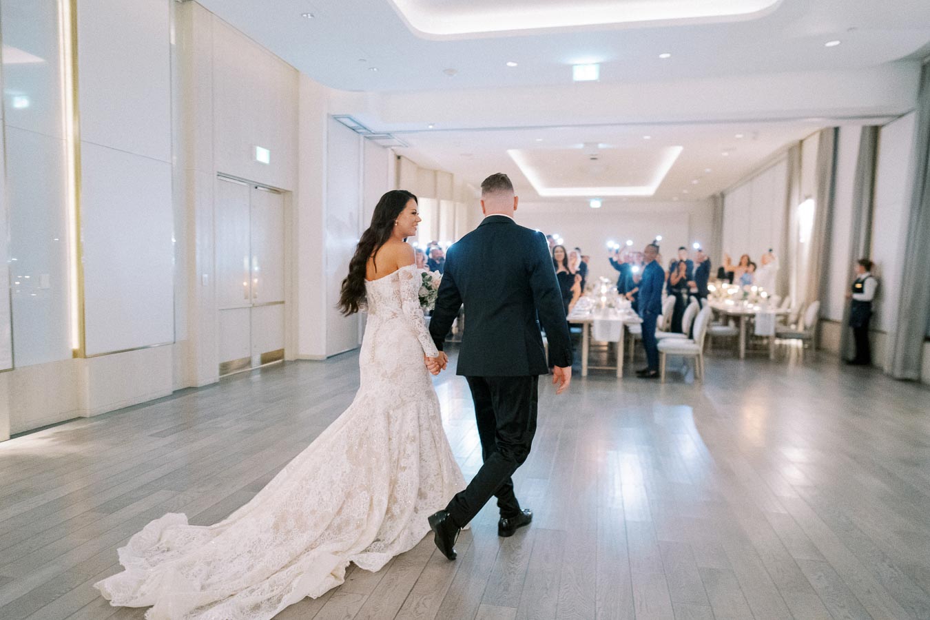 Elegant wedding couple entering a modern reception hall hand in hand, with guests celebrating and photographing the memorable moment.