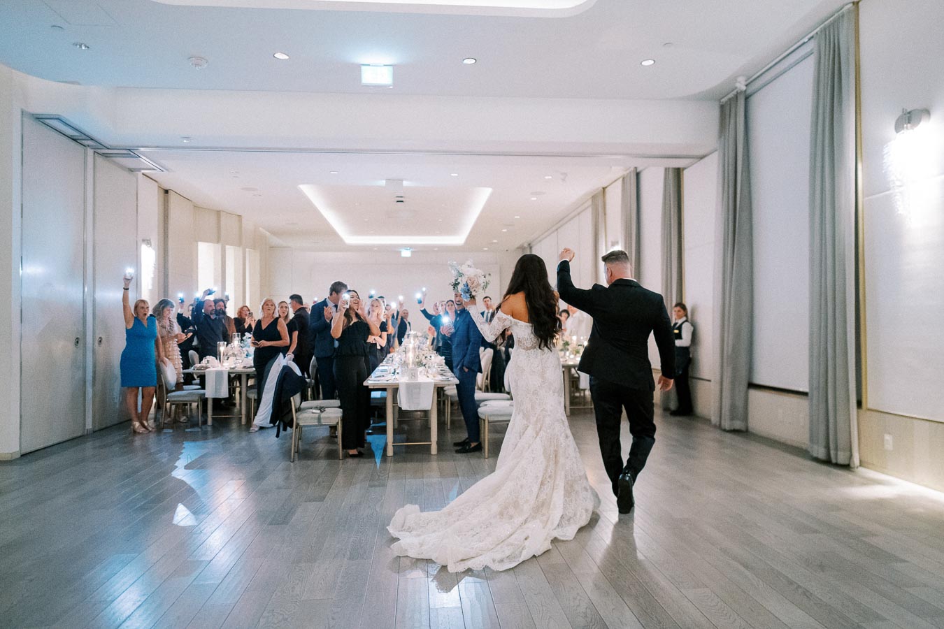 A bride and groom enter a modern wedding reception venue, greeted by cheering guests and phone lights, showcasing an elegant and festive celebration atmosphere.