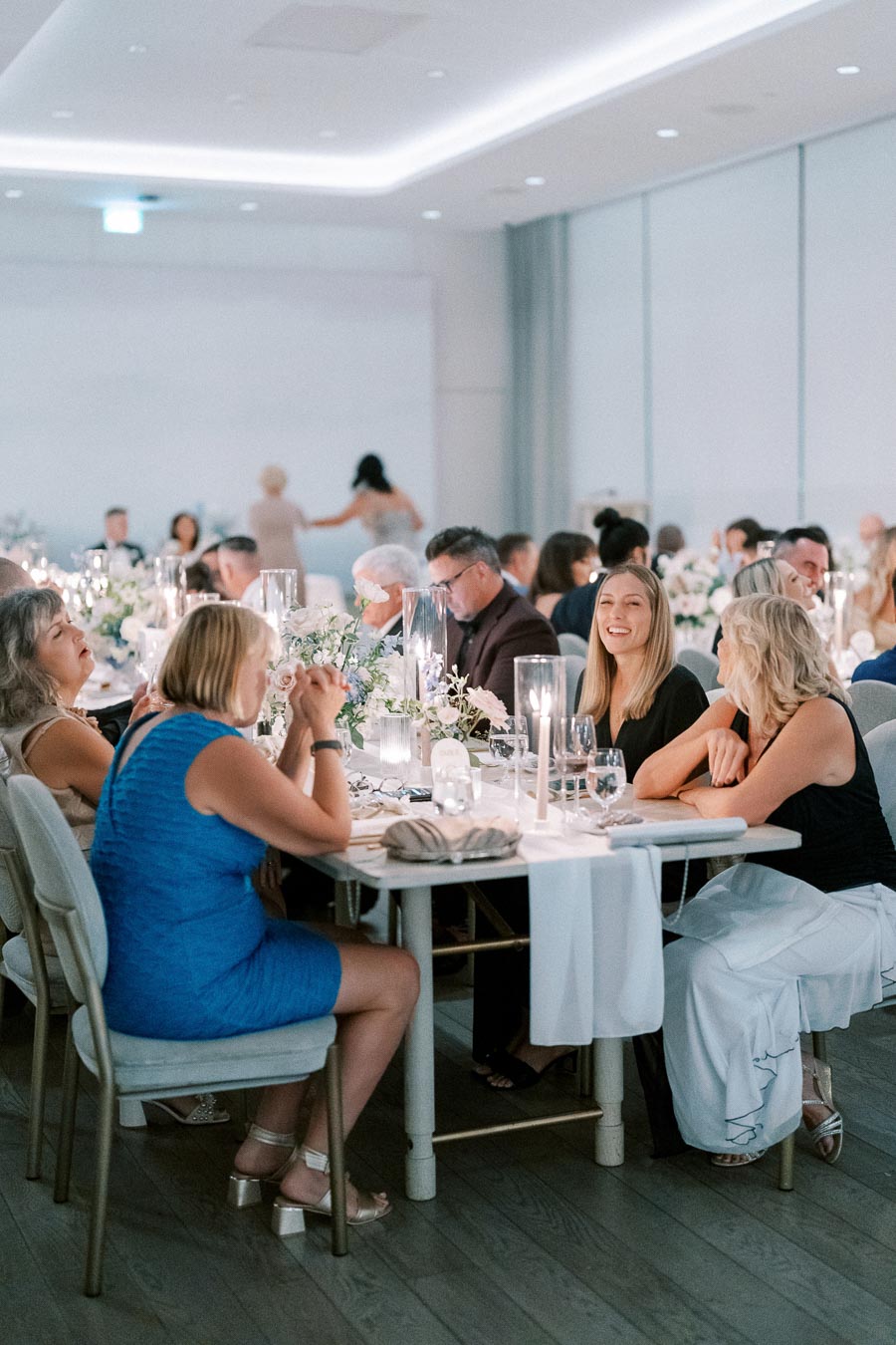 Elegant dinner event with guests seated around a beautifully decorated table, featuring white flowers and candle centerpieces, in a modern, softly lit venue.