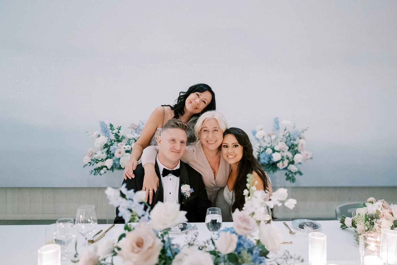 Wedding reception with a group of four smiling attendees, elegantly dressed, seated at a decorated table with floral arrangements, candles, and glassware.