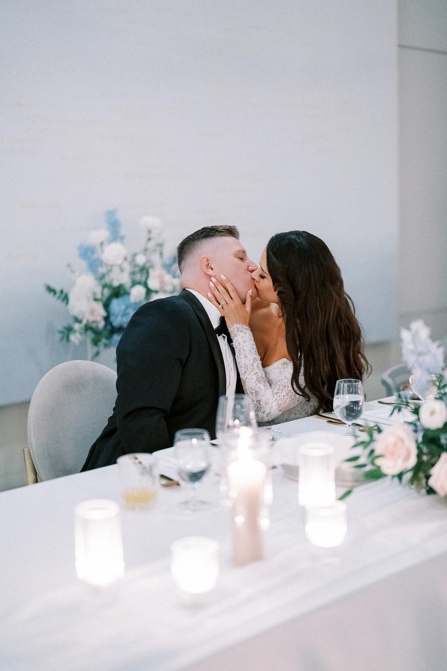 A newlywed couple sharing a romantic kiss at their elegantly decorated wedding reception table, adorned with candles and floral arrangements.