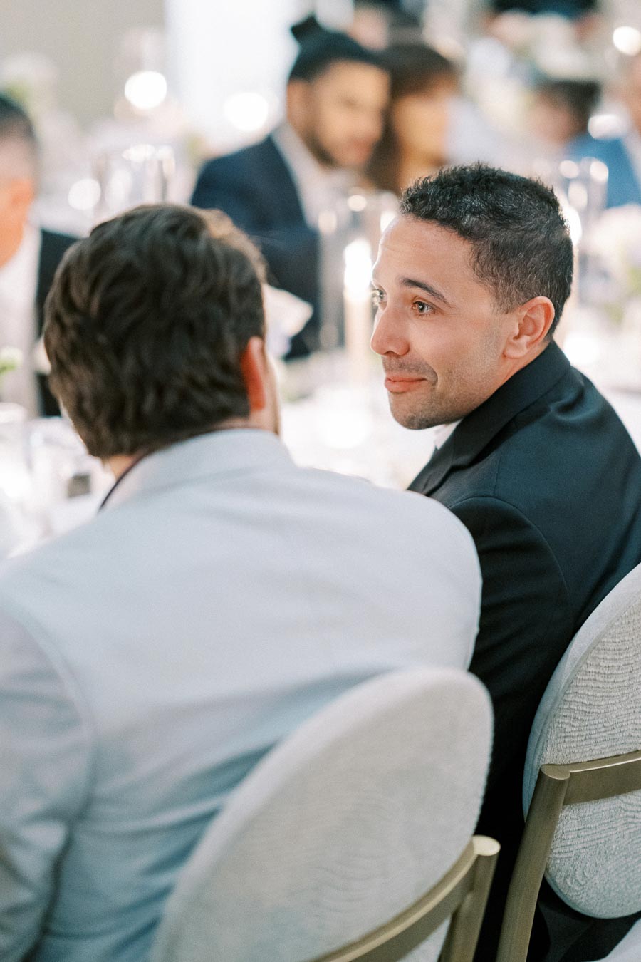 Two men in formal attire sitting at a decorated event table, engaged in conversation, with blurred guests in the background.