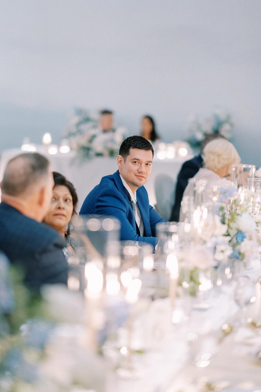 A man in a blue suit sits at a elegantly set wedding reception table, surrounded by other guests with blurred floral arrangements and candles in the background.