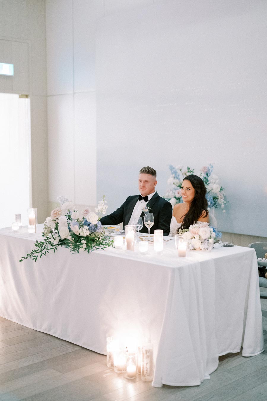 Wedding reception scene with a couple sitting at a beautifully decorated sweetheart table. The table is adorned with white and blue floral arrangements, candles, and elegant tableware. The couple is dressed formally, creating a romantic and sophisticated atmosphere for their special day.
