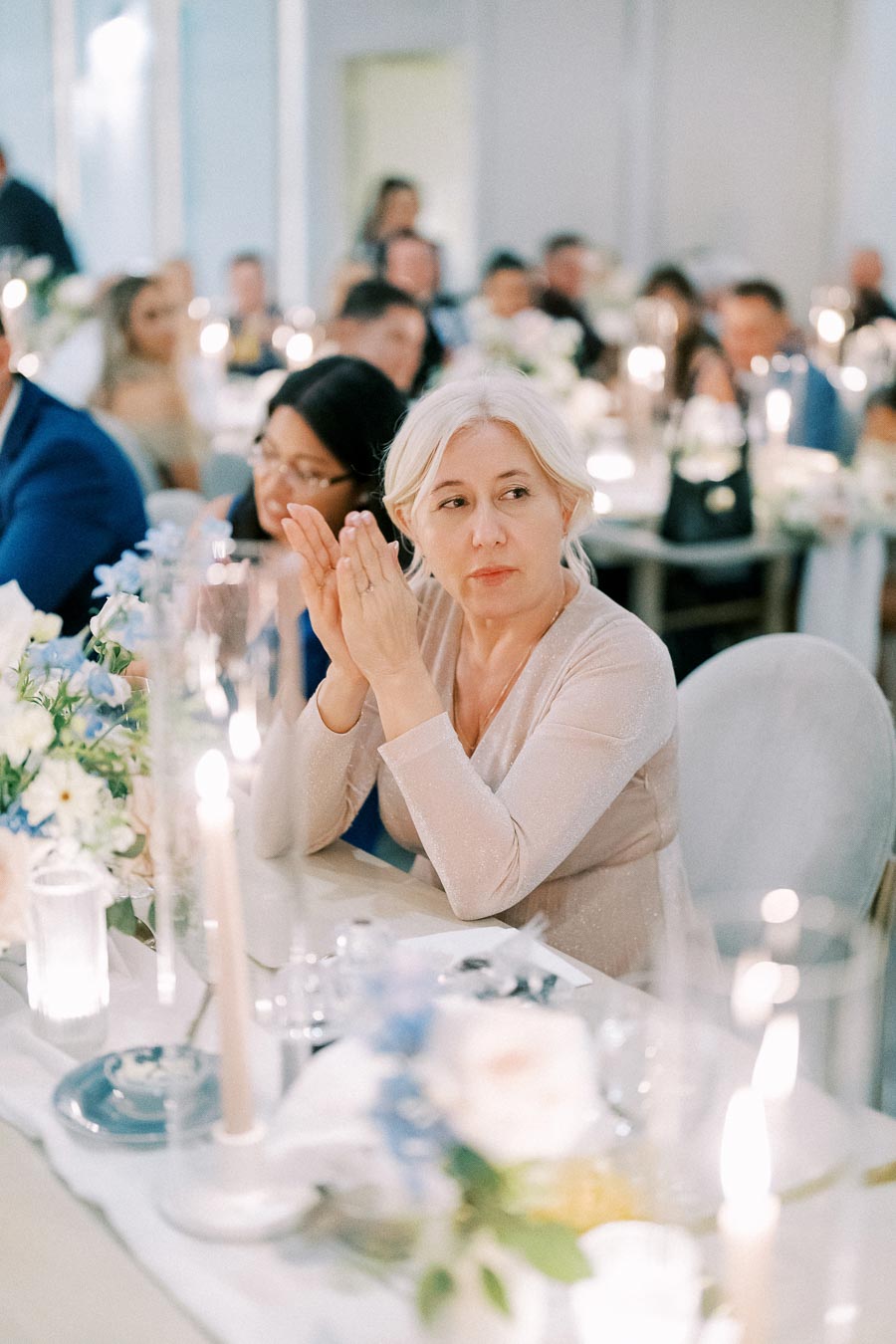 Elderly woman with white hair clapping at a formal event, surrounded by softly lit candles and floral arrangements, with a group of people in the background.