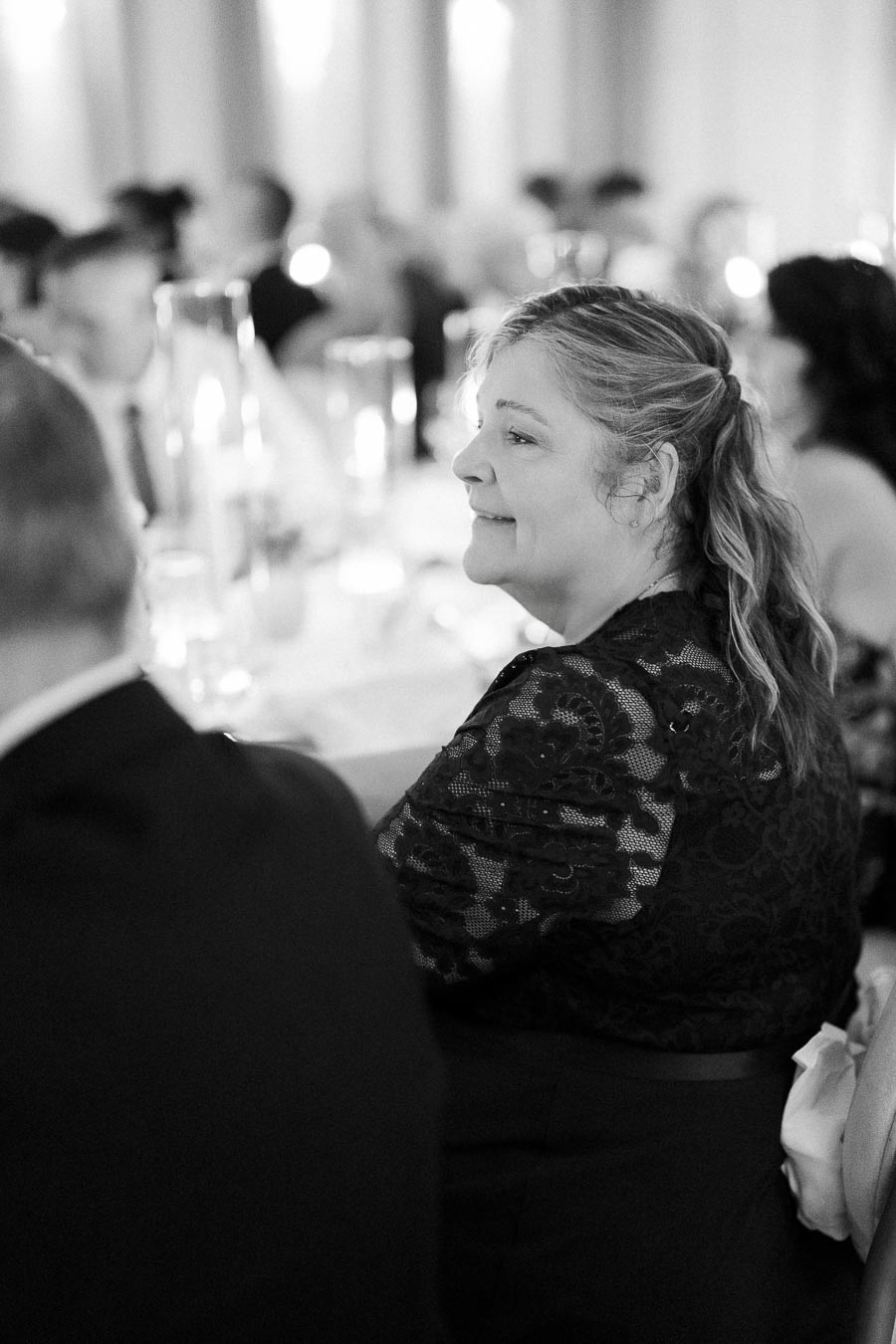 Black and white photo of a woman in elegant attire smiling at a formal event, with blurred guests in the background.