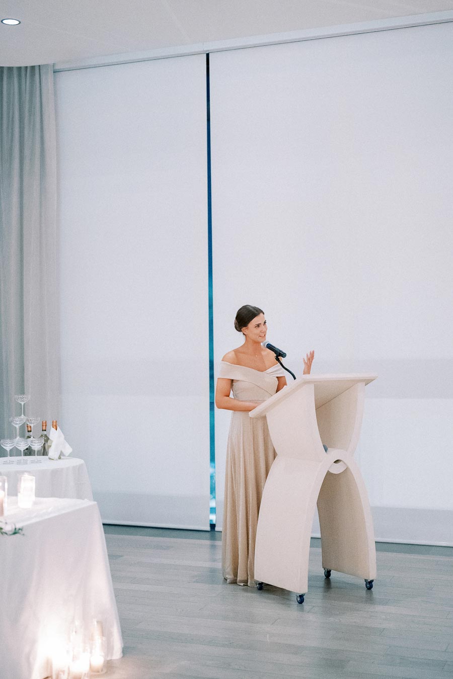 Bridesmaid in elegant off-shoulder gown giving a speech at a wedding reception, standing at a modern white podium with microphones, surrounded by soft lighting and table decor.