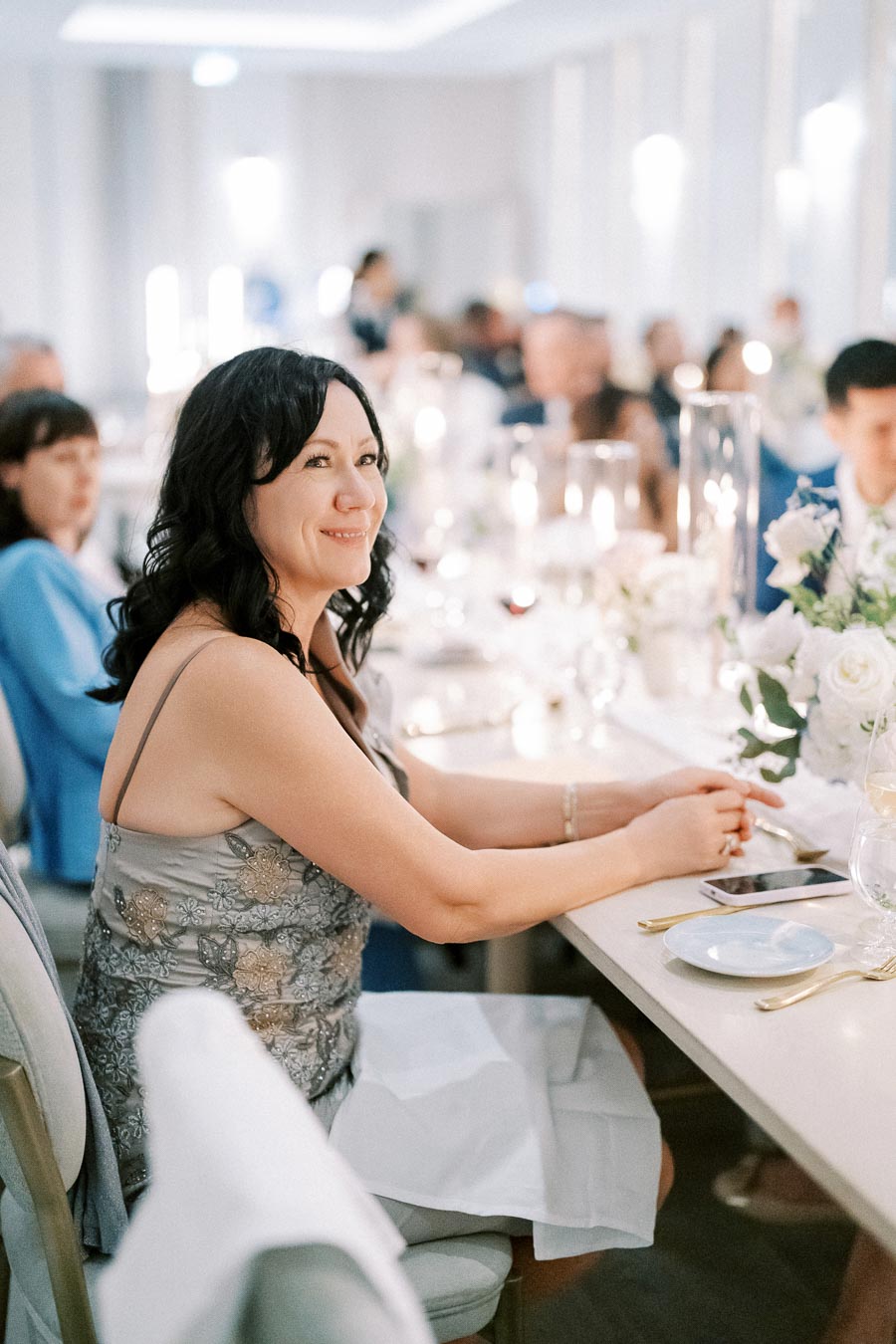 A woman in a floral dress sits smiling at an elegantly decorated event table, adorned with wine glasses and floral centerpieces, during a social gathering.