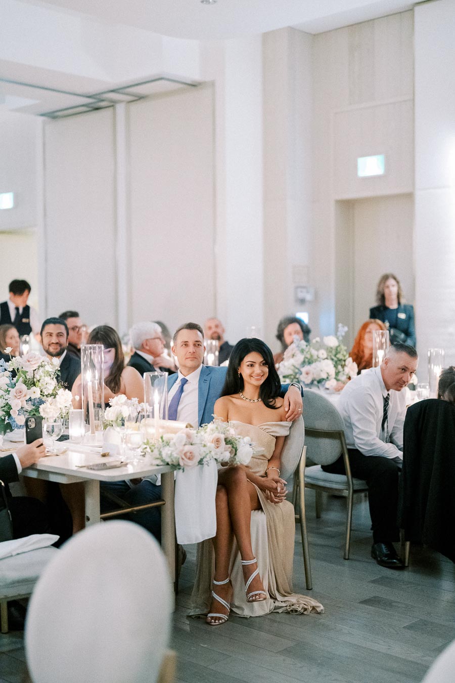 Elegant wedding reception with guests seated at decorated tables, featuring floral centerpieces and candlelight in a modern, bright venue.