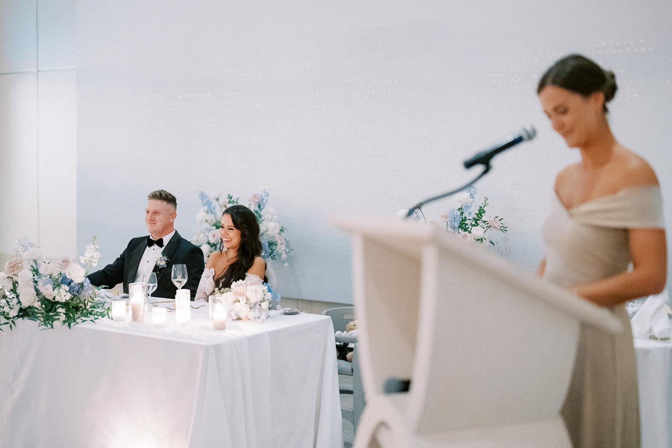 Wedding reception scene with a bride and groom seated at a decorated table, listening to a speech by a woman at a podium, elegant floral arrangements and candles in the foreground.