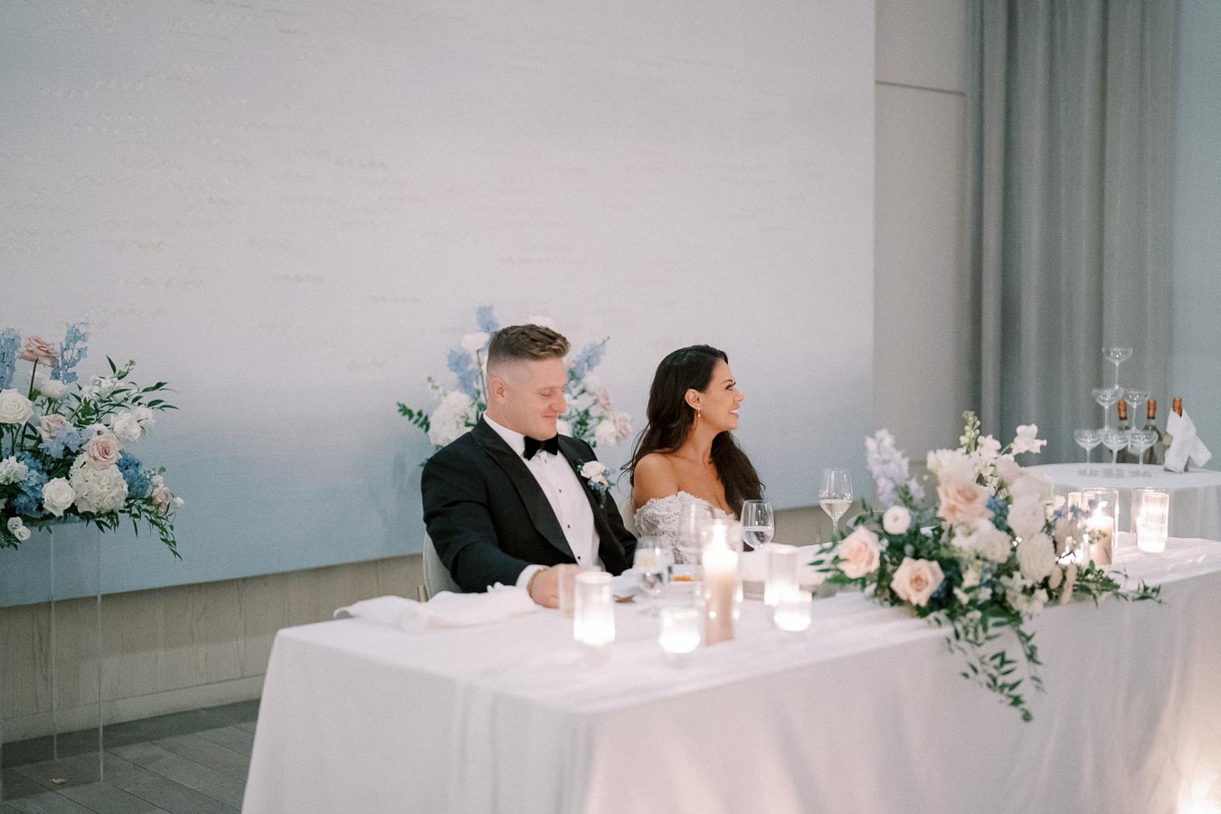Wedding couple sitting at a beautifully decorated reception table with floral arrangements, candles, and wine glasses, in an elegant setting with a soft background.