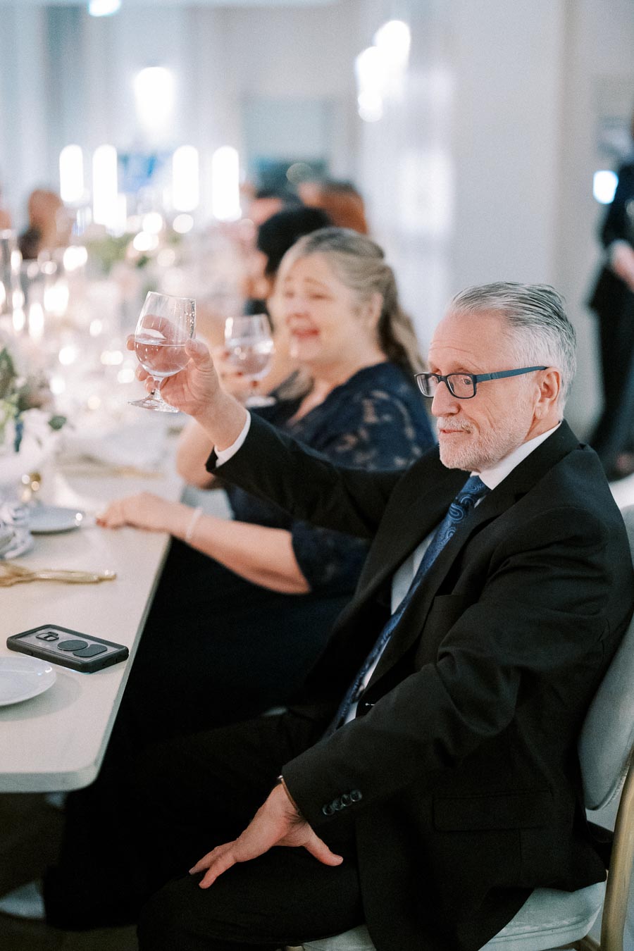 Elderly man in a suit toasting with a wine glass at an elegant dinner party, seated at a beautifully set table with soft lighting and other guests in the background.