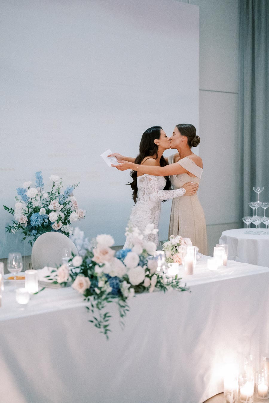 Two brides share a kiss at their wedding reception, surrounded by elegant floral arrangements and candlelit tables.