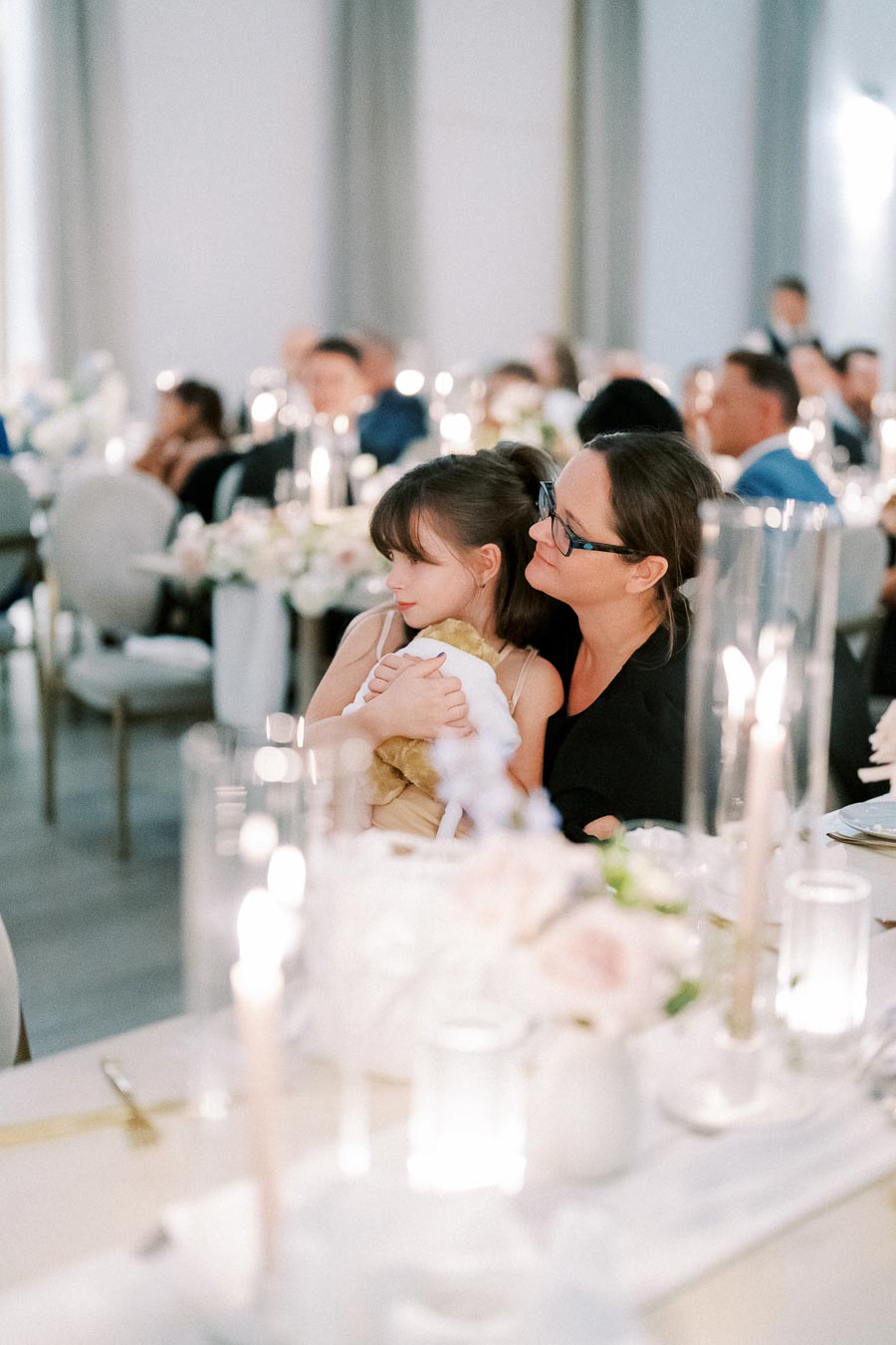 A woman and child seated at an elegant wedding reception, surrounded by softly lit candles and floral arrangements, creating a warm and intimate atmosphere.
