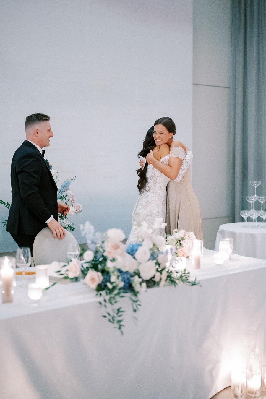 A bride and bridesmaid share a joyful embrace at a wedding reception, with a groomsman standing nearby. The setting features elegant floral arrangements and candles on a table, creating a romantic atmosphere.