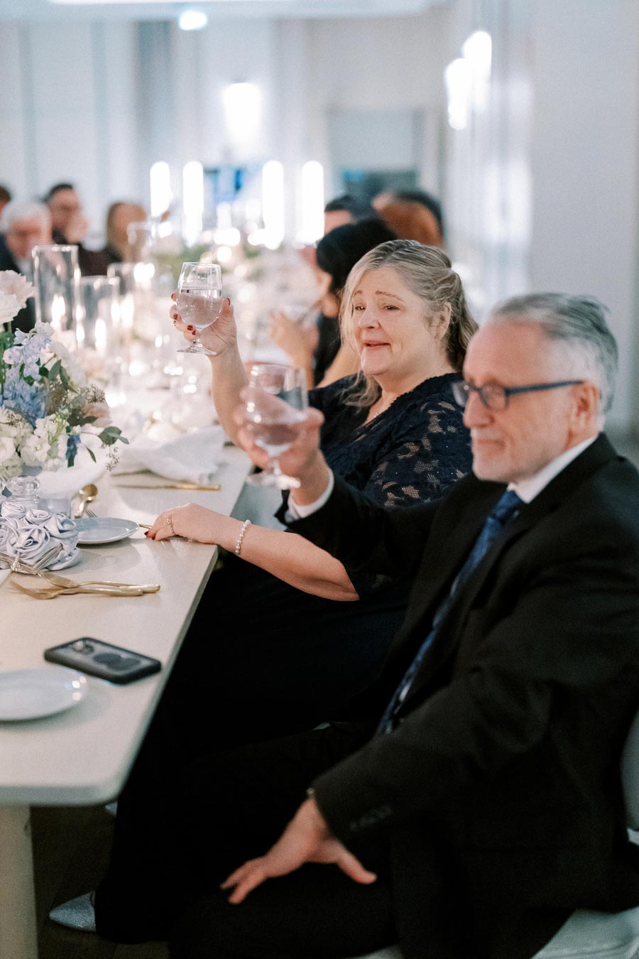 A joyful gathering at a formal event, featuring elegantly dressed guests raising glasses for a toast around a beautifully decorated dinner table with floral arrangements and candles.