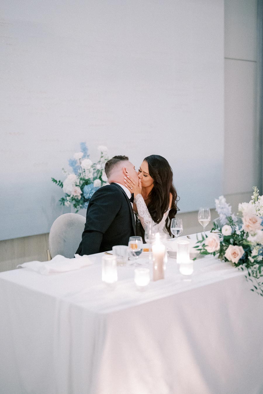 Elegant wedding reception scene with a couple kissing at a beautifully decorated table adorned with flowers and candles.
