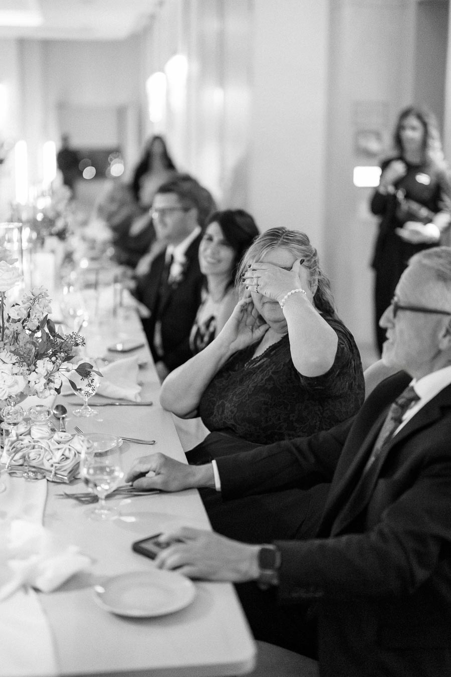 Black and white photo of elegantly dressed people sitting at a decorated dining table during a formal event, with one person playfully covering their face, laughing and engaging with nearby guests.