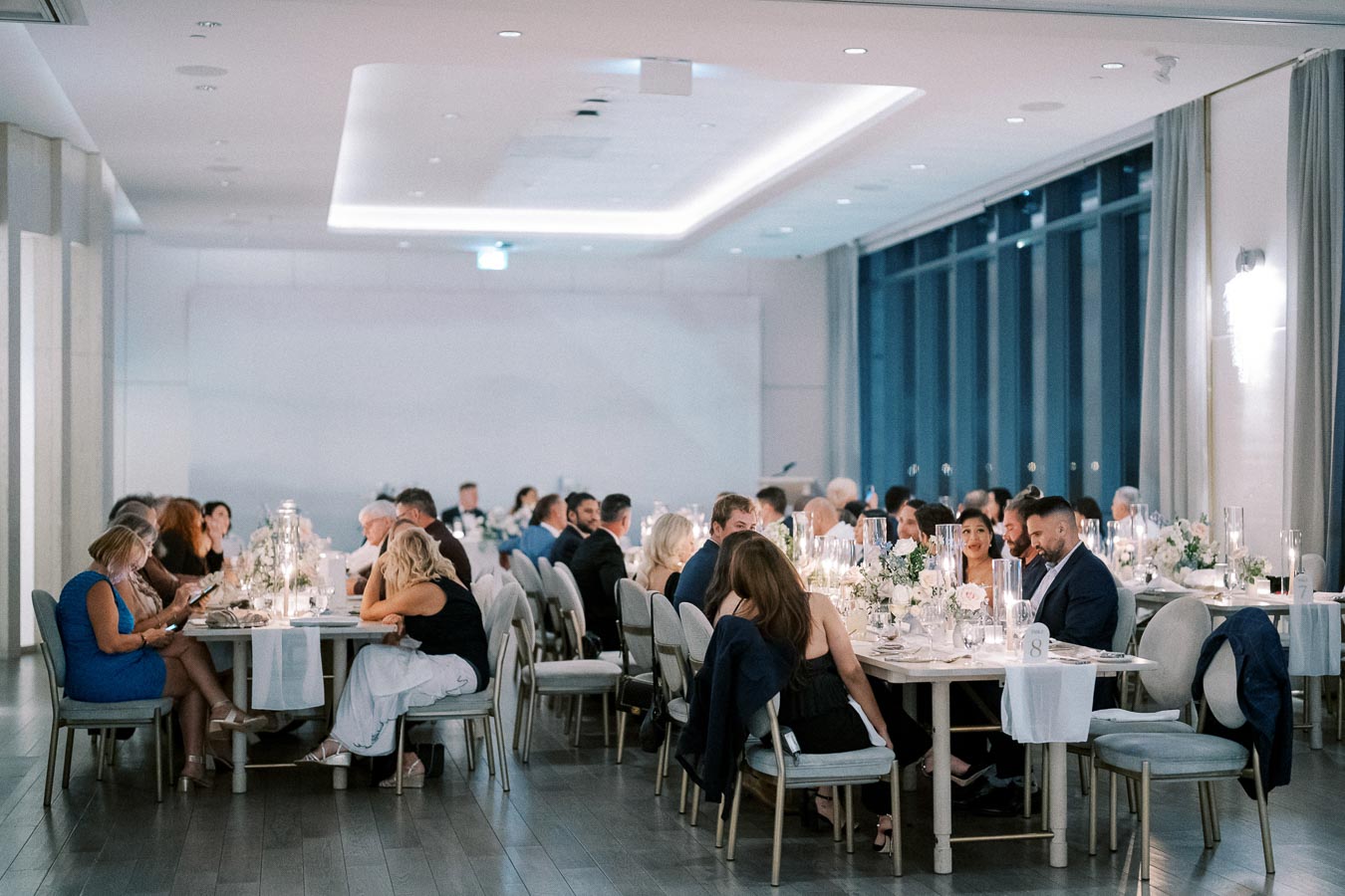 Elegant wedding reception with guests seated at decorated tables in a modern, softly lit venue.