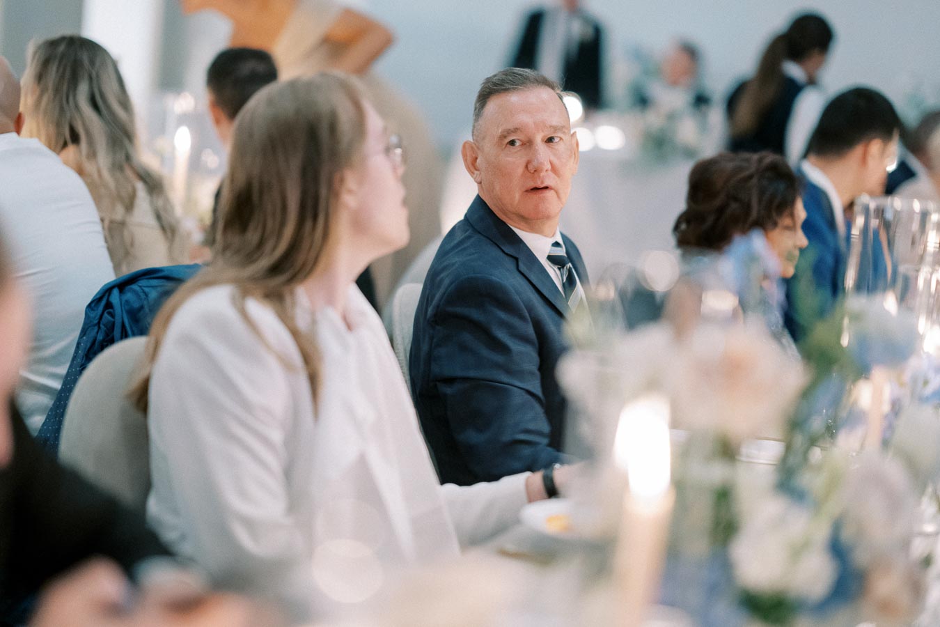 Guests seated at a formal event, engaged in conversation, with elegantly set tables in a softly lit venue.