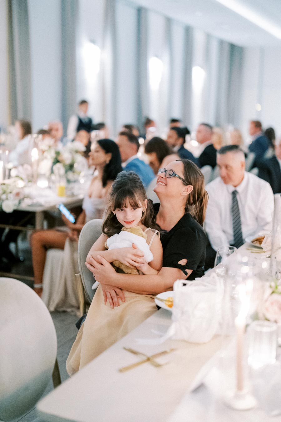 A joyful mother and daughter sit together at a wedding reception, surrounded by elegantly dressed guests and a beautifully set table with floral centerpieces and glowing candles.