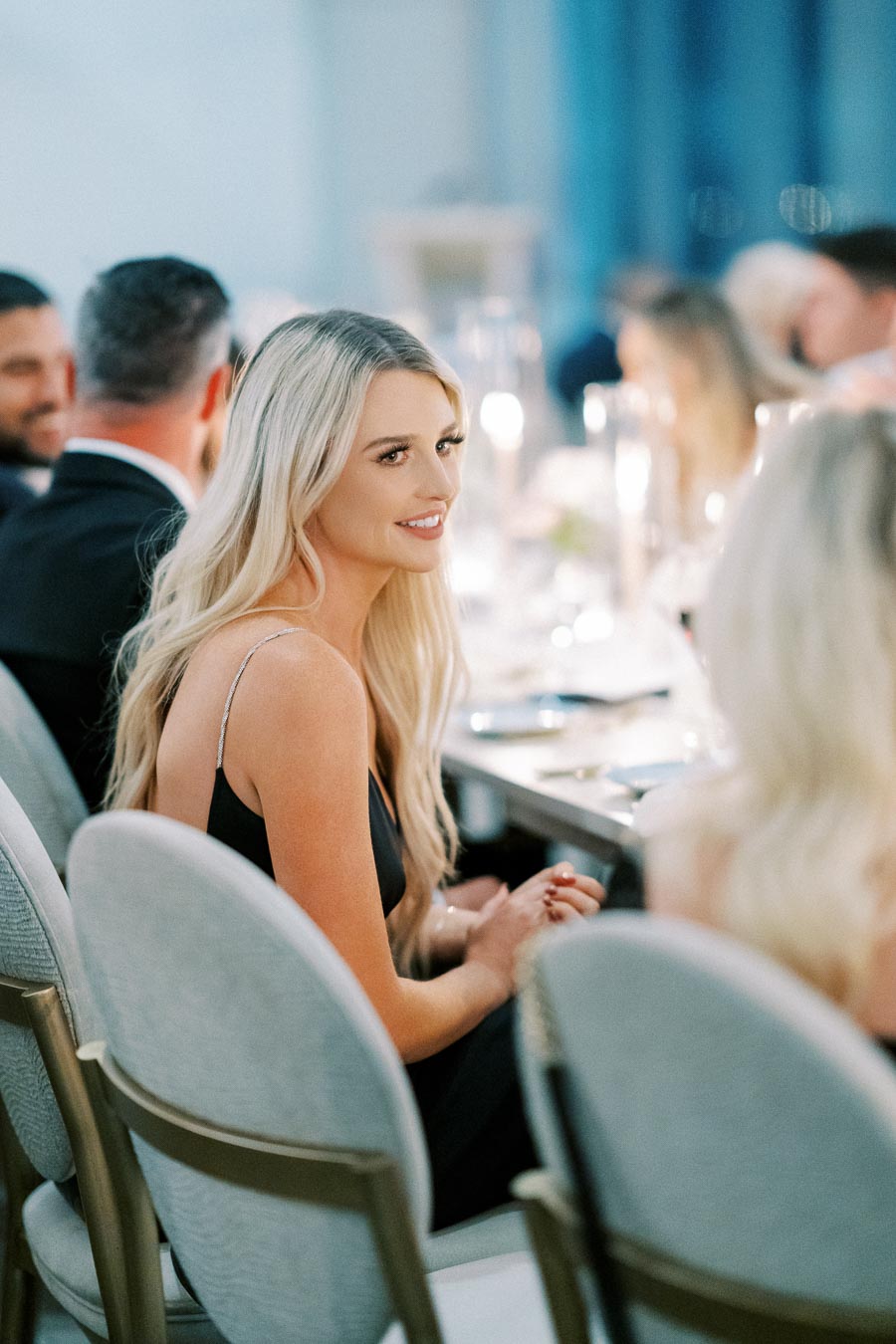 Smiling woman with long blonde hair seated at an elegant dinner event, surrounded by other guests in formal attire.