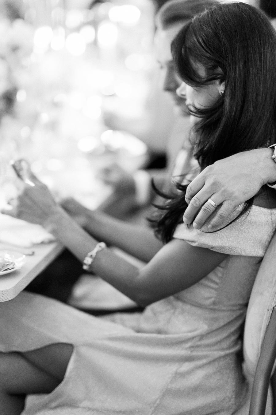 Monochrome photo of a couple seated at an elegantly set table, with a man's hand gently resting on a woman's shoulder, creating an intimate and romantic atmosphere.