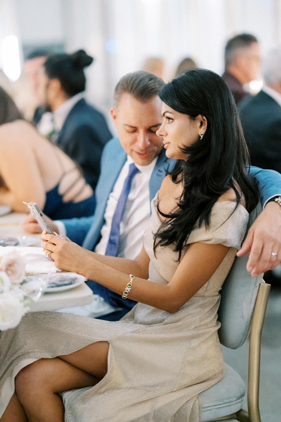 Elegant couple sitting at a formal event, with a woman in a shimmering dress using a smartphone while a man in a blue suit is seated next to her.