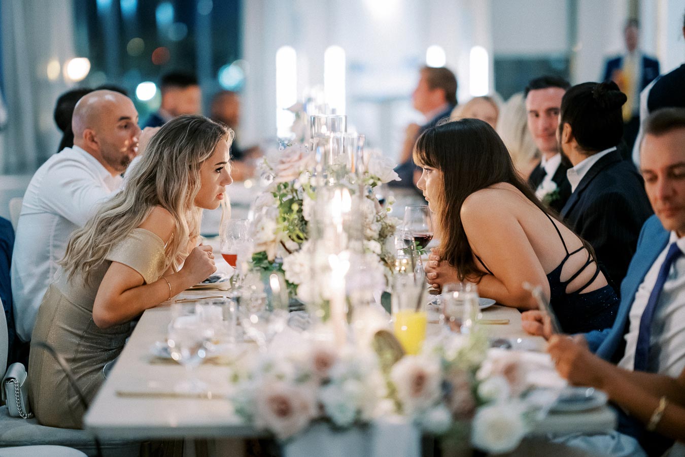 Guests engaged in conversation at a decorated dining table during a formal event, with floral centerpieces and ambient lighting.