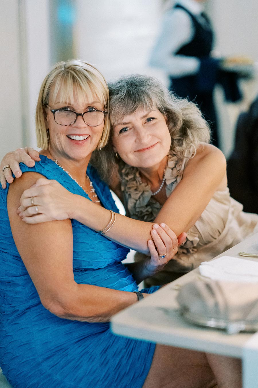 Two smiling women sitting together at a table, with one wearing a blue dress and the other a beige outfit, showcasing a warm embrace and joyful atmosphere.