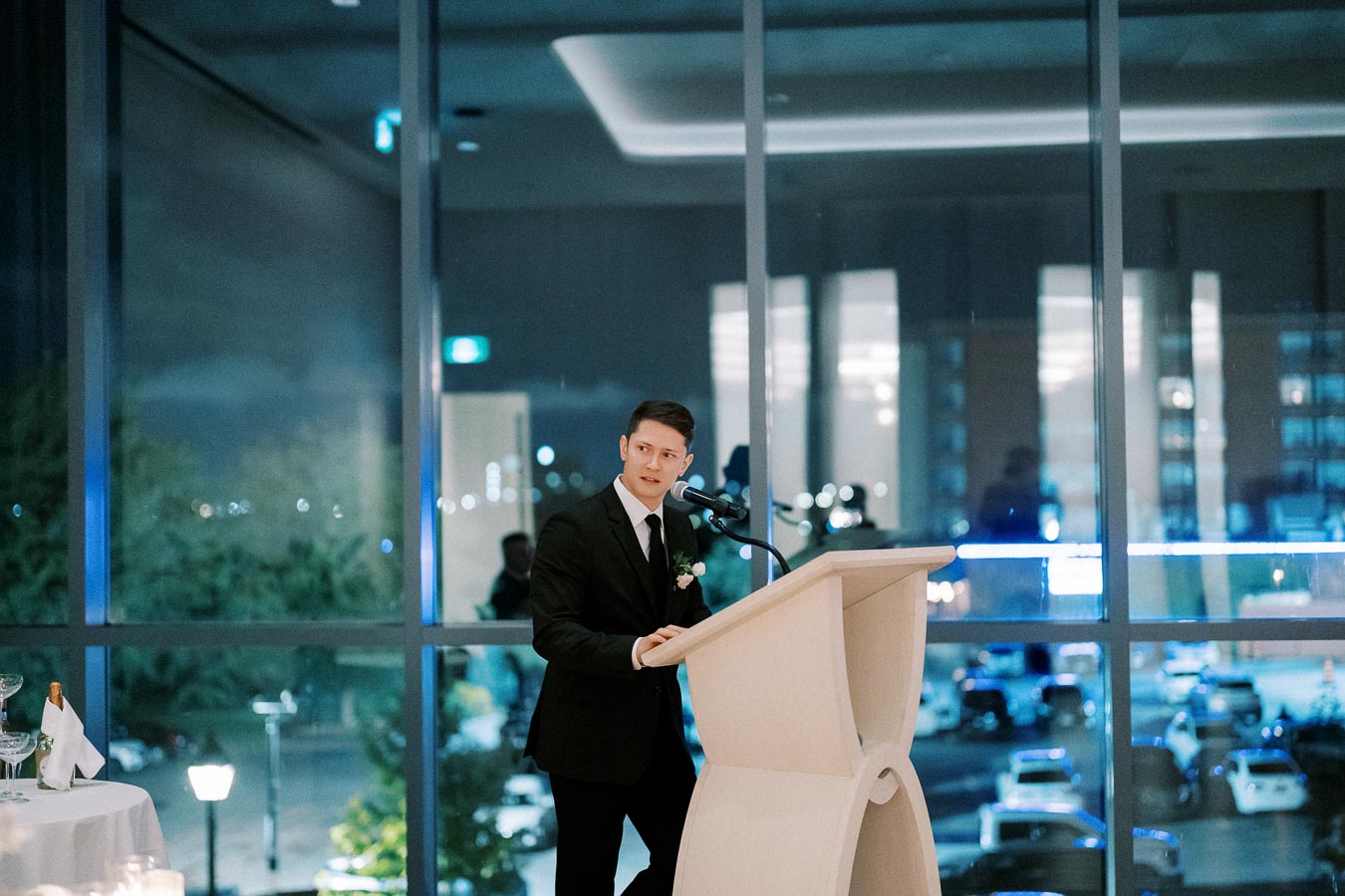 A man in a suit giving a speech at a podium in a modern venue with large windows overlooking a cityscape at night.
